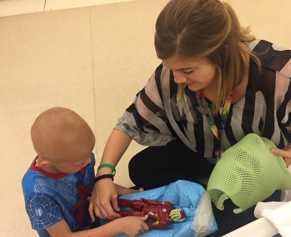 Специалист по работе с детьми в центре NMCPC и пациент-ребенок накладывают фиксирующую маску на игрушечную фигурку